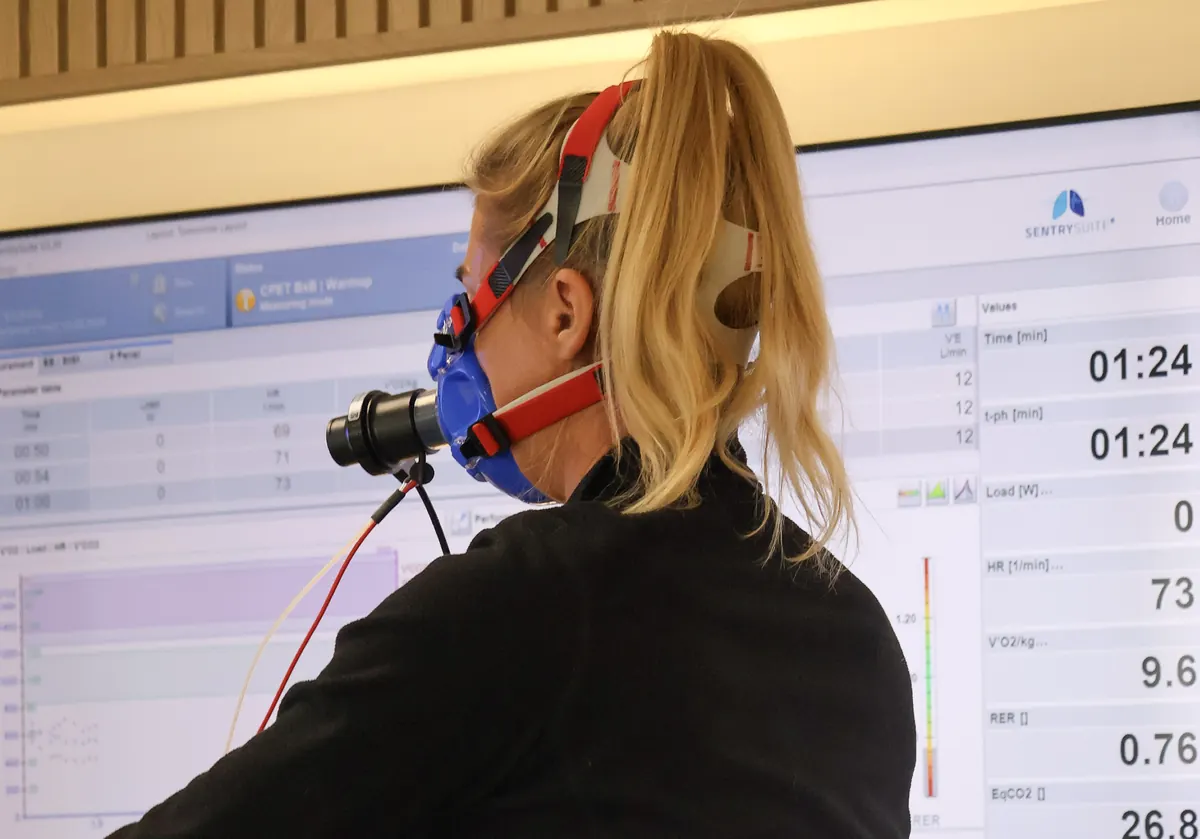 A client on the exercise bike wearing a breathing mask during a heart and lung test, with live readings on the screen behind her